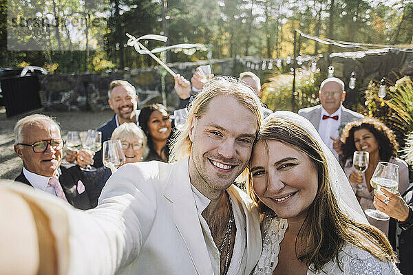 Porträt eines glücklichen frisch vermählten Paares  das am Hochzeitstag ein Selfie mit Gästen im Hintergrund macht