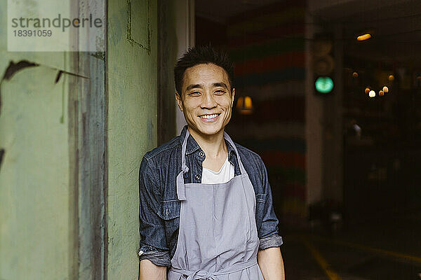 Porträt eines lächelnden Restaurantbesitzers in Schürze  der an der Wand lehnt