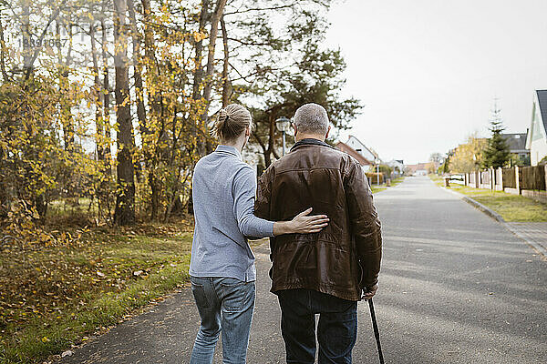 Rückansicht eines Hausmeisters  der einen älteren Mann im Ruhestand unterstützt  der mit einem Stock auf der Straße geht