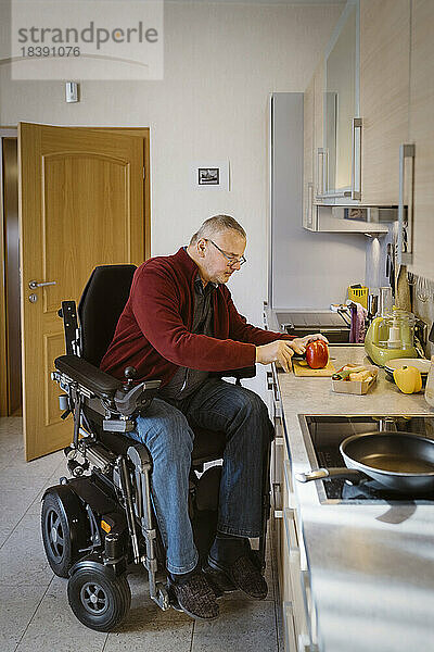 Rentner mit Behinderung sitzt im motorisierten Rollstuhl und schneidet Paprika an der Küchentheke in voller Länge