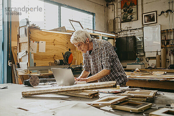 Fokussierter älterer männlicher Tischler bei der Arbeit an einem Laptop in einer Reparaturwerkstatt