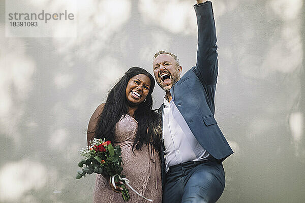 Fröhlich reifen Bräutigam schreien von jungen Braut mit Blumenstrauß gegen die Wand bei der Hochzeit