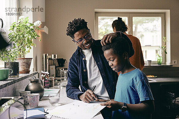 Lächelnder Vater streichelt das Haar seines Sohnes  der zu Hause Hausaufgaben macht