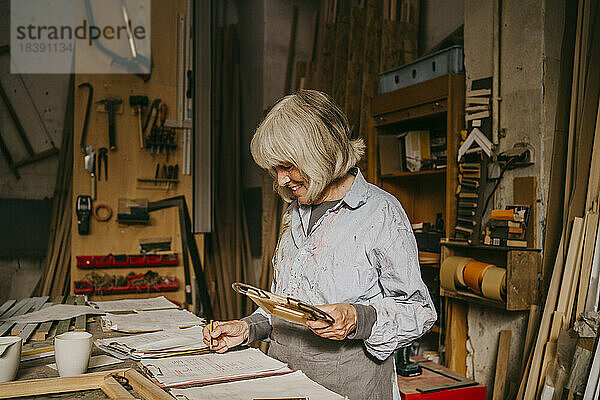Lächelnder älterer weiblicher Schreiner  der einen Rahmen hält  während er in der Werkstatt auf einem Klemmbrett schreibt