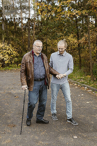 Junger männlicher Hausmeister in voller Länge  der einen älteren Mann im Ruhestand unterstützt  der mit einem Stock auf der Straße geht