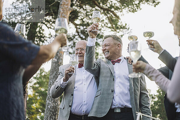 Glückliches frisch verheiratetes schwules Paar erhebt seine Weingläser mit Familie und Freunden bei der Hochzeit