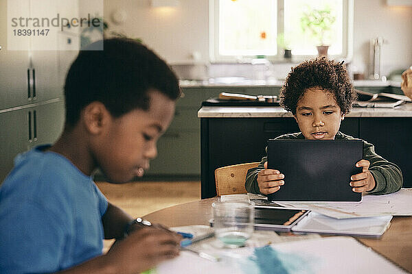 Junge benutzt digitales Tablet  während sein Bruder zu Hause in einem Buch malt