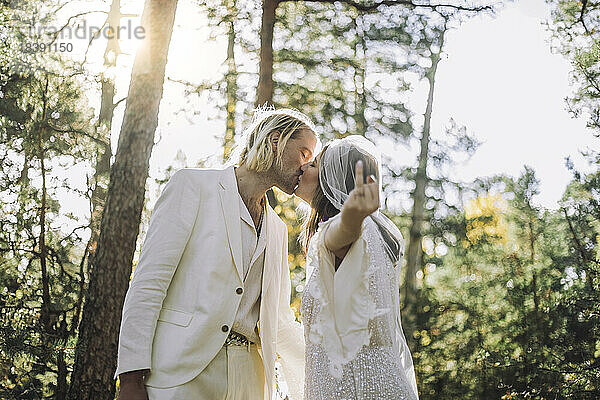 Braut zeigt Ringfinger und küsst Bräutigam auf den Mund im Wald während der Hochzeit