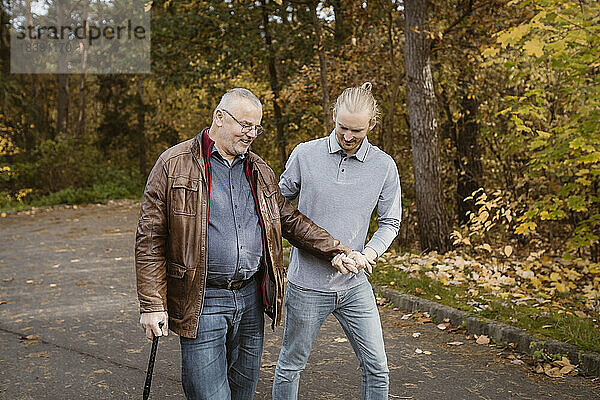 Junger männlicher Pfleger unterstützt einen älteren Mann im Ruhestand  der mit einem Stock auf der Straße geht