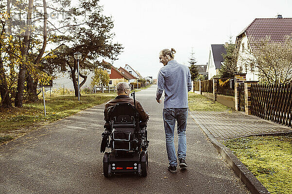 Rückansicht eines älteren Mannes mit Behinderung im motorisierten Rollstuhl  der von einer jungen Betreuerin auf der Straße begleitet wird