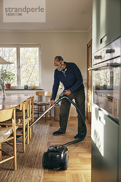 Älterer Mann reinigt Teppich mit Staubsauger im Stehen zu Hause