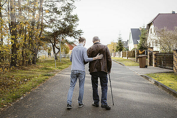 Rückansicht eines Hausmeisters in voller Länge  der einen älteren Mann im Ruhestand unterstützt  der mit einem Stock auf der Straße geht