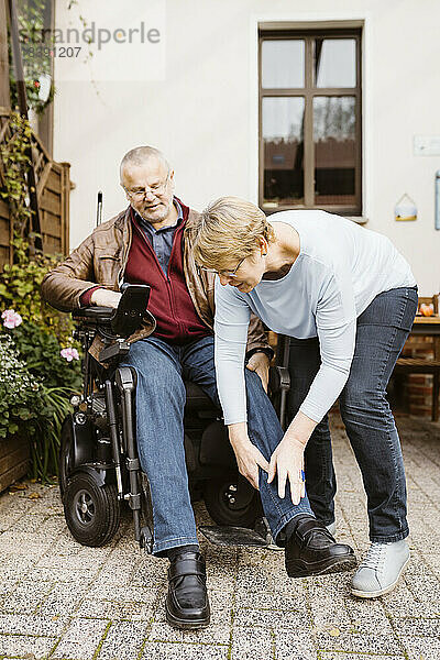 Ältere Frau in voller Länge  die einem Mann mit Behinderung hilft  der in einem motorisierten Rollstuhl im Hinterhof sitzt