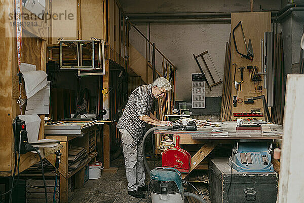 Seitenansicht eines älteren männlichen Schreiners  der in einer Werkstatt stehend an einem Laptop arbeitet