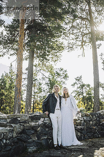 Porträt eines frisch verheirateten Paares  das sich an eine Stützmauer vor einem Wald lehnt