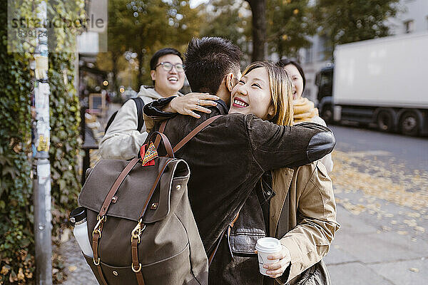 Glückliche junge Frau umarmt männlichen Freund  der einen Rucksack trägt  während sie auf dem Bürgersteig steht