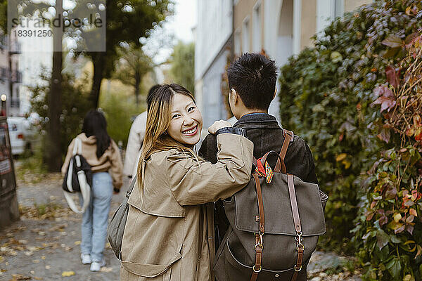 Porträt einer glücklichen Frau  die über die Schulter schaut  während sie mit einem männlichen Freund auf dem Gehweg spazieren geht
