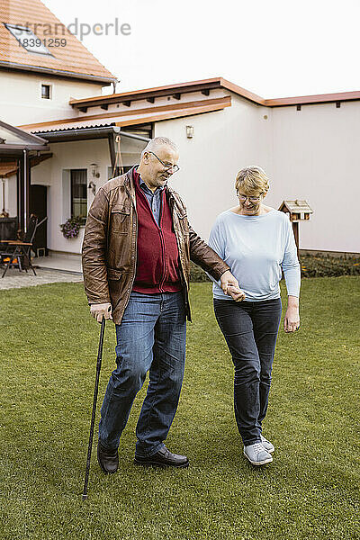 Älterer Mann  der die Hand einer Frau hält  während er mit einem Stock auf einer Wiese im Hinterhof geht