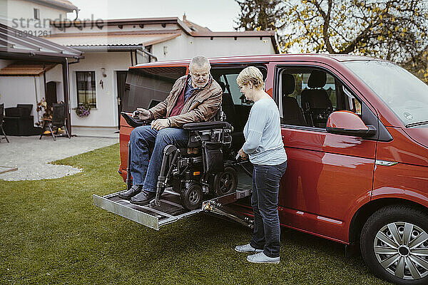 Ältere Frau in voller Länge  die eine Fernbedienung hält  während sie einem Mann mit einer Behinderung in einem motorisierten Rollstuhl hilft  auszusteigen