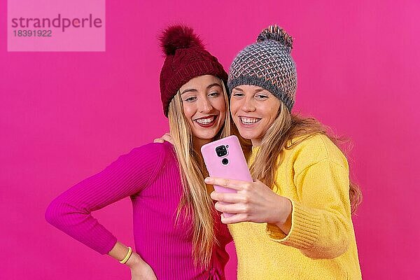 Zwei junge blonde kaukasische Frauen mit einem Telefon vor einem rosa Hintergrund  Selfie lächelnd