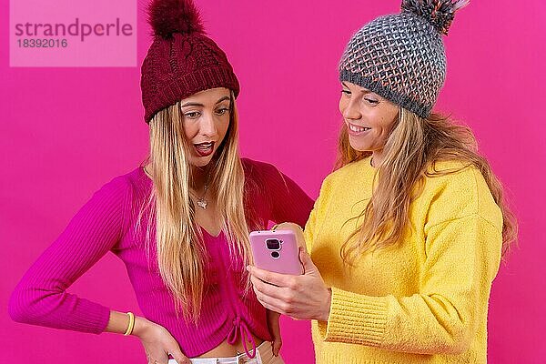 Zwei junge blonde kaukasische Frauen mit einem Telefon vor einem rosa Hintergrund  Blick auf das Internet