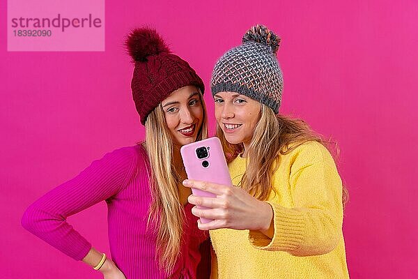 Zwei junge blonde kaukasische Frauen mit einem Telefon vor einem rosa Hintergrund  Selfie lächelnd