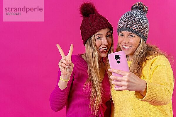 Zwei junge blonde kaukasische Frauen mit einem Telefon vor einem rosa Hintergrund  Selfie machen die ok Geste