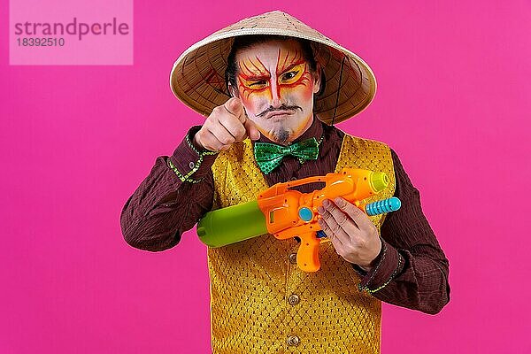 Clown mit weißer Gesichtsschminke auf rosa Hintergrund  der sich mit Spielzeugpistolen vergnügt