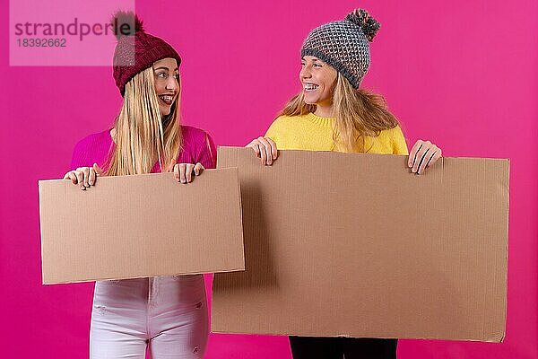 Zwei junge blonde kaukasische Frauen halten ein Schild vor einem rosa Hintergrund  Studioaufnahme