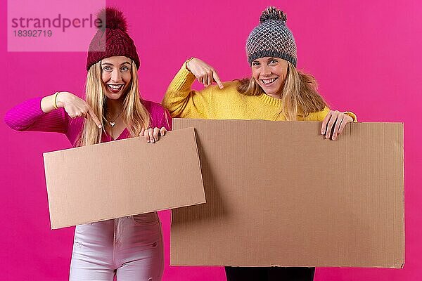 Zwei junge blonde Frauen  die auf ein Schild zeigen  vor einem rosa Hintergrund