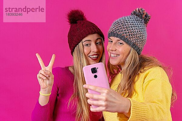 Zwei junge blonde kaukasische Frauen mit einem Telefon vor einem rosa Hintergrund  Selfie machen die ok Geste