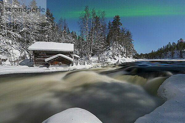 Einsame Hütte im verschneiten Wald mit Blick auf die Myllikoski-Stromschnellen des zugefrorenen Flusses unter Nordlicht  Ruka Kuusamo  Lappland  Finnland  Europa
