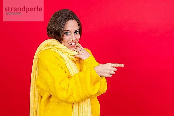 Attraktive Frau  die lächelnd mit dem Finger auf den Kopierraum auf rotem Hintergrund zeigt  gelber Wollpullover