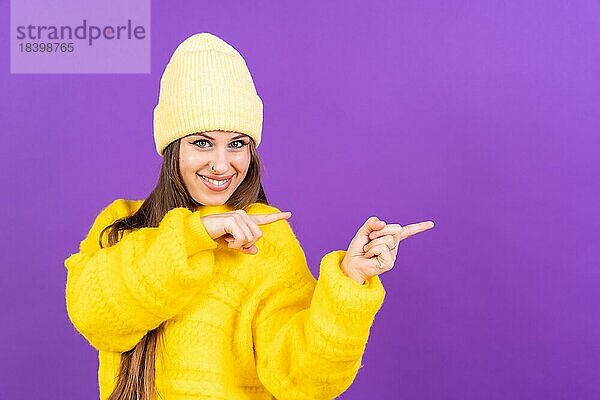 Attraktive Frau  die lächelnd mit den Fingern auf den Kopierraum auf lila Hintergrund zeigt  gelber Wollpullover