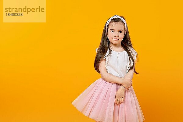Ein süßes kleines Mädchen steht in einem gelb getönten Studio und schaut unschuldig in die Kamera. Ihr langes Haar und ihr modisch geschmücktes Kleid spiegeln ihre Kindheit wider  während sie diesen Moment der Schönheit festhält