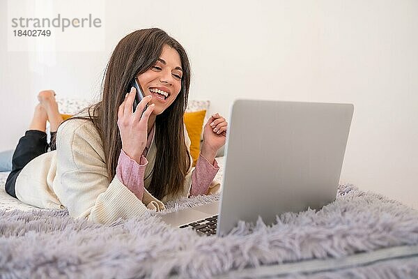 Frau arbeitet mit Laptop im Bett  Telearbeit  lächelnd am Telefon