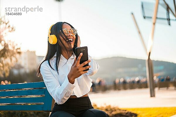 Afroamerikanische Frau hört Musik und lächelt mit Kopfhörern im Sommer bei Sonnenuntergang in der Stadt