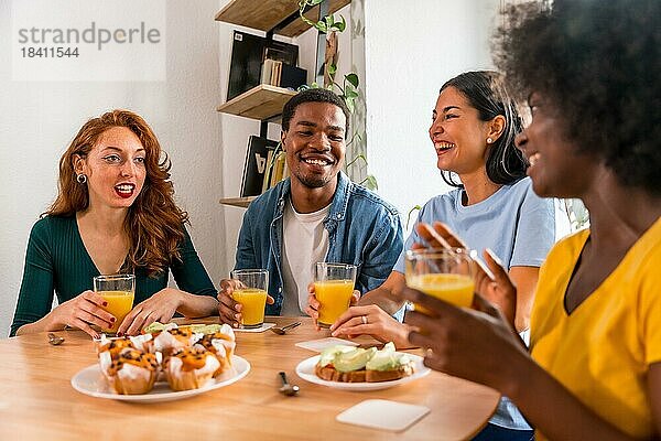 Lächelnde multiethnische Freunde beim Frühstück mit Orangensaft und Muffins zu Hause