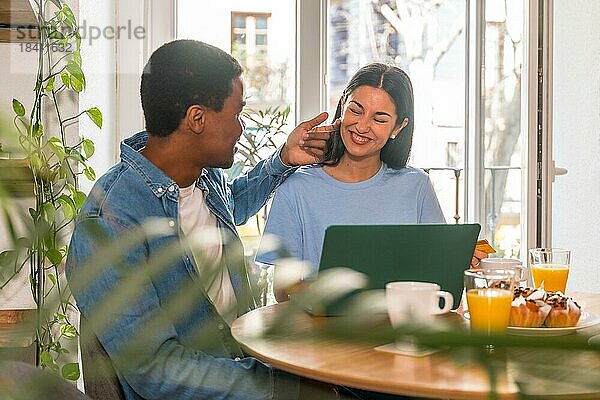 Verliebtes multiethnisches Paar  das beim Frühstück am Fenster einen Online Einkauf über den Computer tätigt