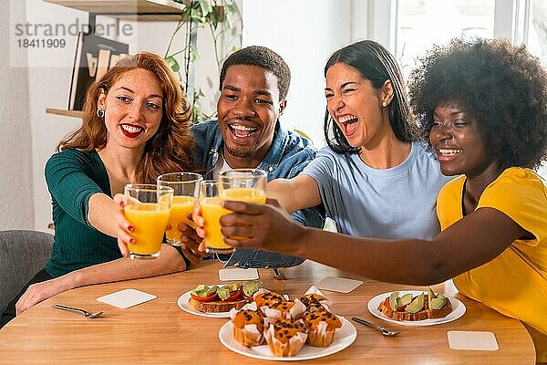 Multiethnische Freunde stoßen bei einem Frühstück mit Orangensaft und Muffins zu Hause an