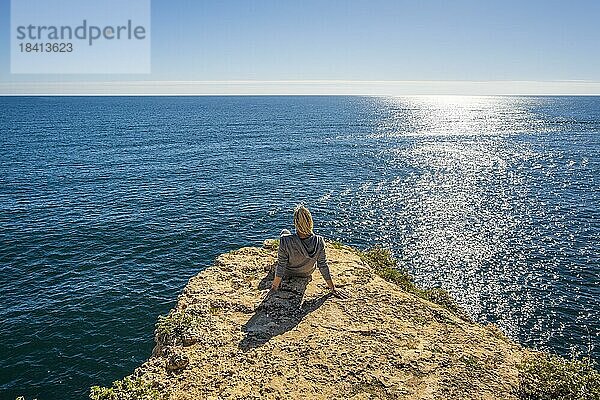 Rückenansicht einer jungen Frau  die auf den Klippen sitzt und die Schönheit des Atlantischen Ozeans am Strand von Marinha in der Algarve  Portugal  genießt  Europa