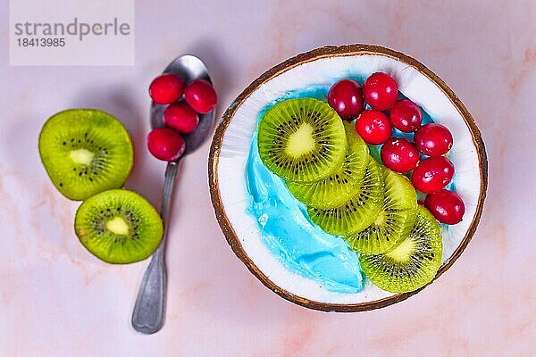 Draufsicht auf eine FruchtSmoothieSchale in einer Kokosnussschale  die natürlich mit blauem Spirulina Pulver gefärbt und mit Cranberries und Kiwis garniert ist