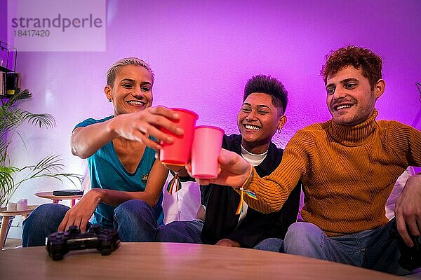 Gruppe junger Freunde  die zusammen auf dem Sofa zu Hause Videospiele spielen und mit farbigen Gläsern anstoßen  junge Millennials