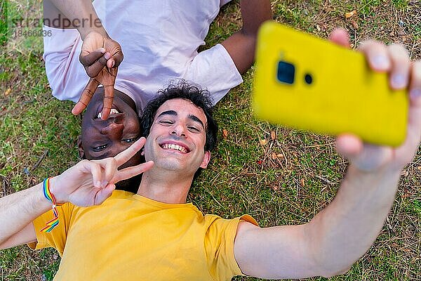 Ein paar multiethnische Männer in einem Park  lgbt Konzept  auf dem Gras liegend  die ein Selfie mit einem gelben Handy machen