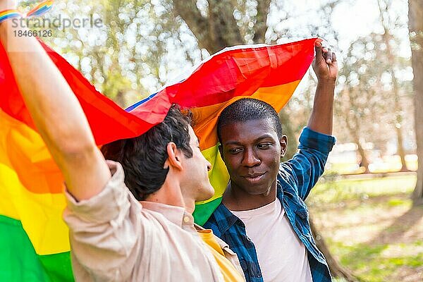 Multiethnisches schwules männliches Paar hält lächelnd eine Regenbogenflagge mit dem Symbol der Homosexualität