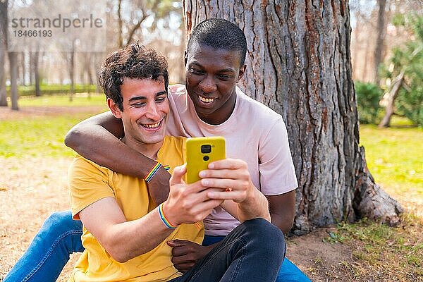 Ein paar multiethnische Männer in einem Park  lgbt Konzept  sitzen neben einem Baum und haben Spaß mit einem gelben Handy