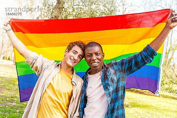 Porträt eines multiethnischen schwulen Paares  das bei Sonnenuntergang eine Regenbogenflagge mit dem Symbol der Homosexualität hält