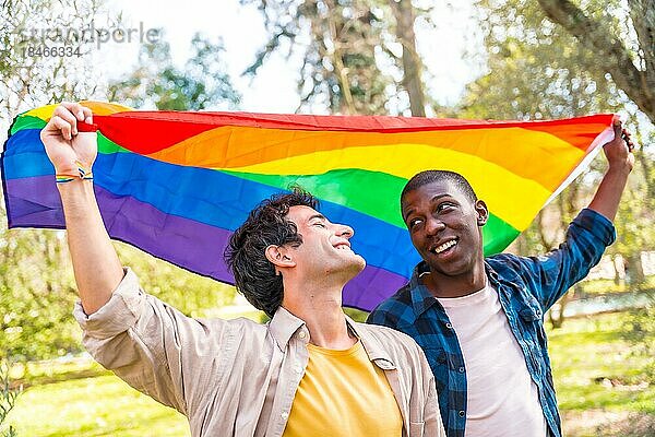 Multiethnisches schwules männliches Paar hält lächelnd eine Regenbogenflagge mit dem Symbol der Homosexualität