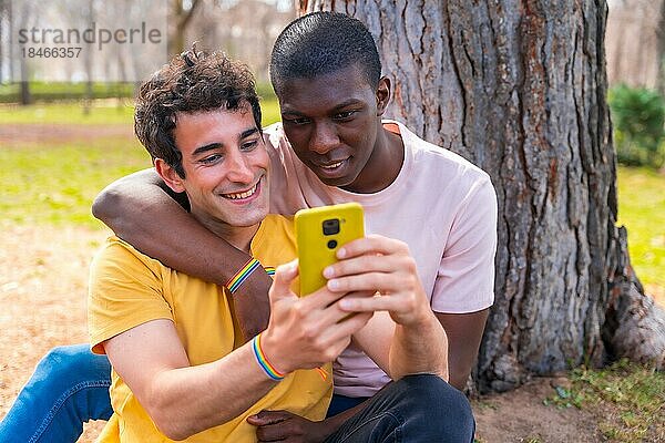 Ein paar multiethnische Männer in einem Park  lgbt Konzept  sitzen neben einem Baum und haben Spaß mit einem gelben Handy