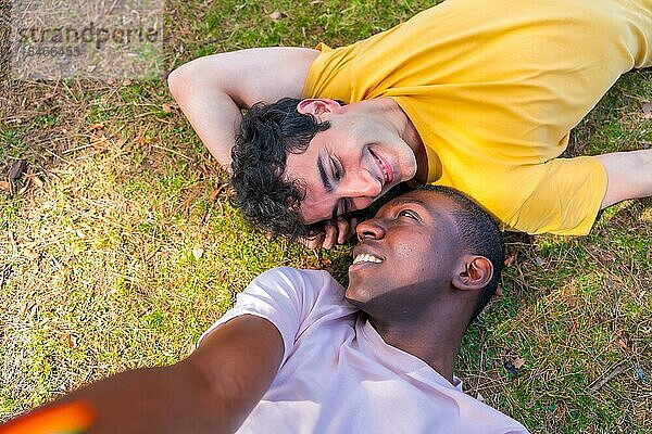 Porträt eines Paares multiethnischer Männer in einem Park  Igbt Konzept  die auf dem Gras liegen und ein Selfie machen